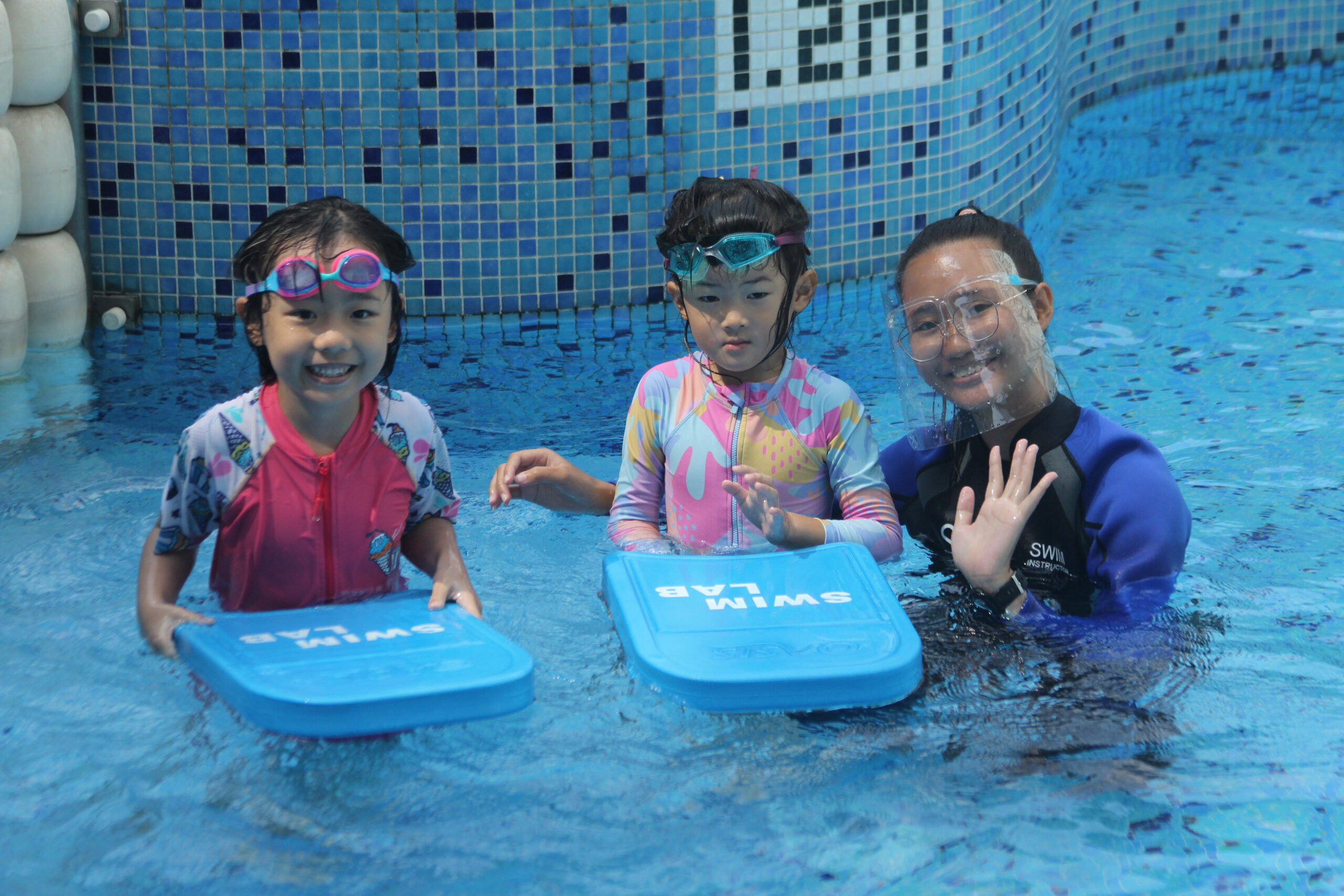 Two children holding kickboards and a swim instructor offering guidance in a pool, capturing an engaging moment of a kids' swimming lesson.