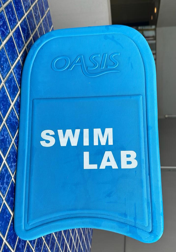 A Swim Lab Kickboard resting on the edge of a swimming pool, a specialized swimming aid designed to focus on lower body strength and kicking skills during swim lessons.