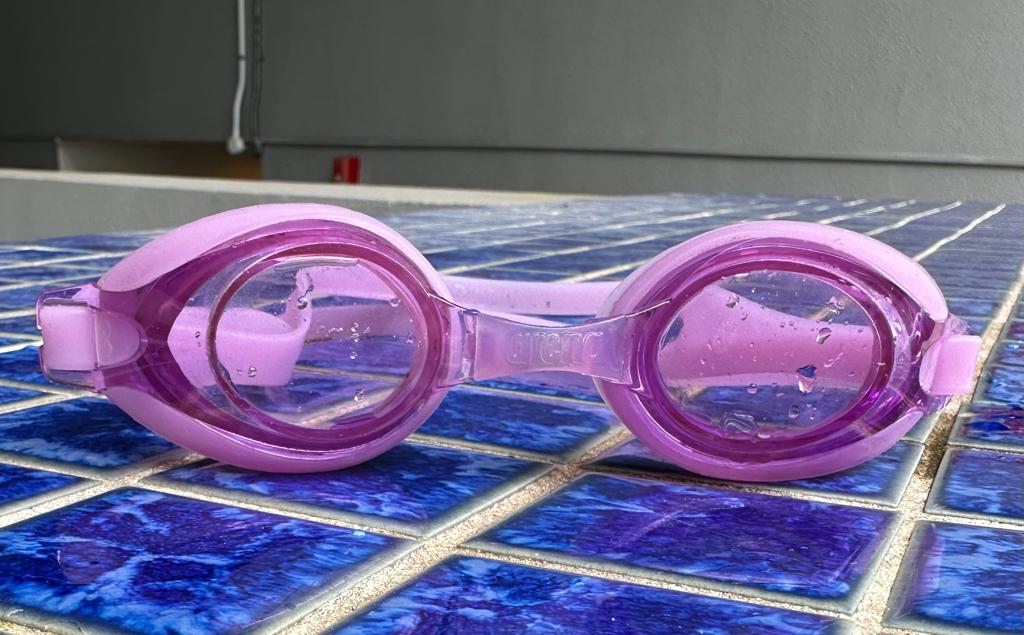 A pair of clear lens swimming goggles displayed poolside, ready to provide unobstructed underwater vision for kids in a swimming class.