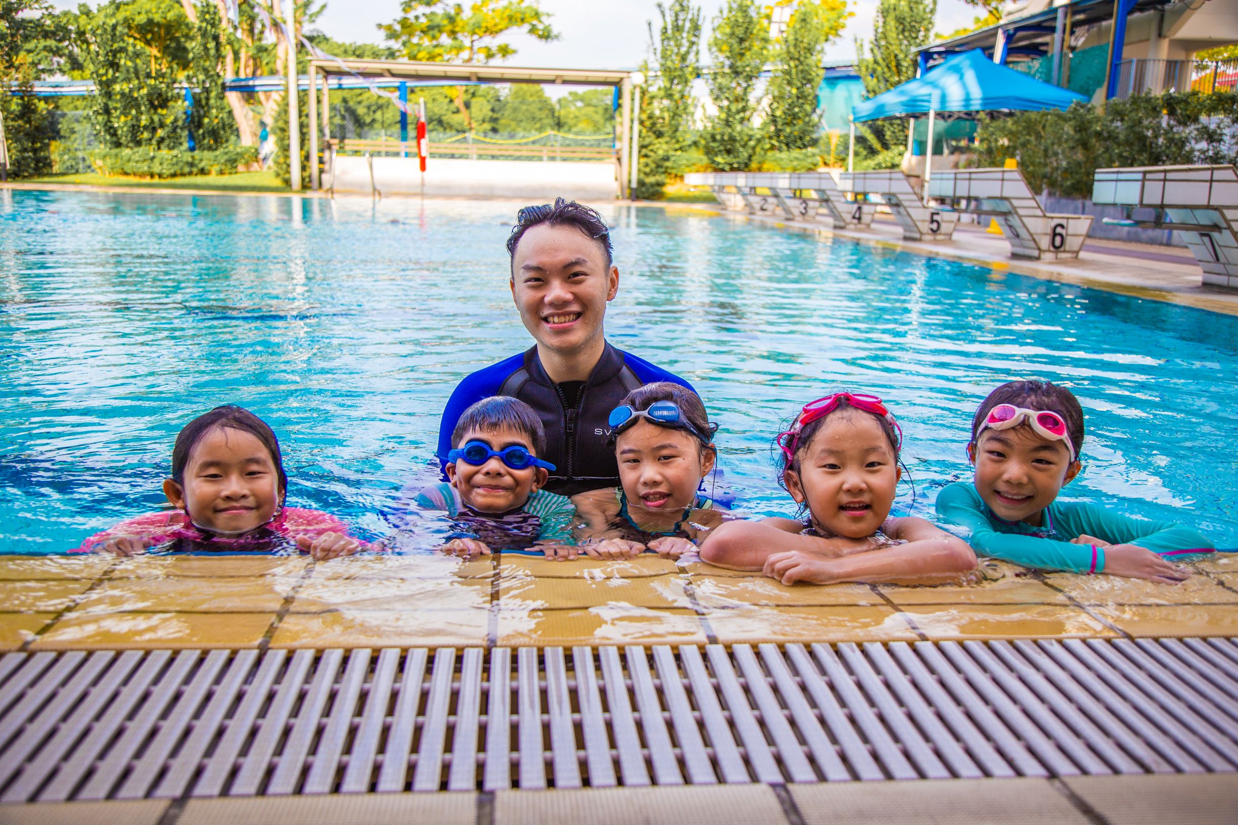 Group of kids with their swim coach, smiling and posing in the clear waters of Hwa Chong swimming pool.
