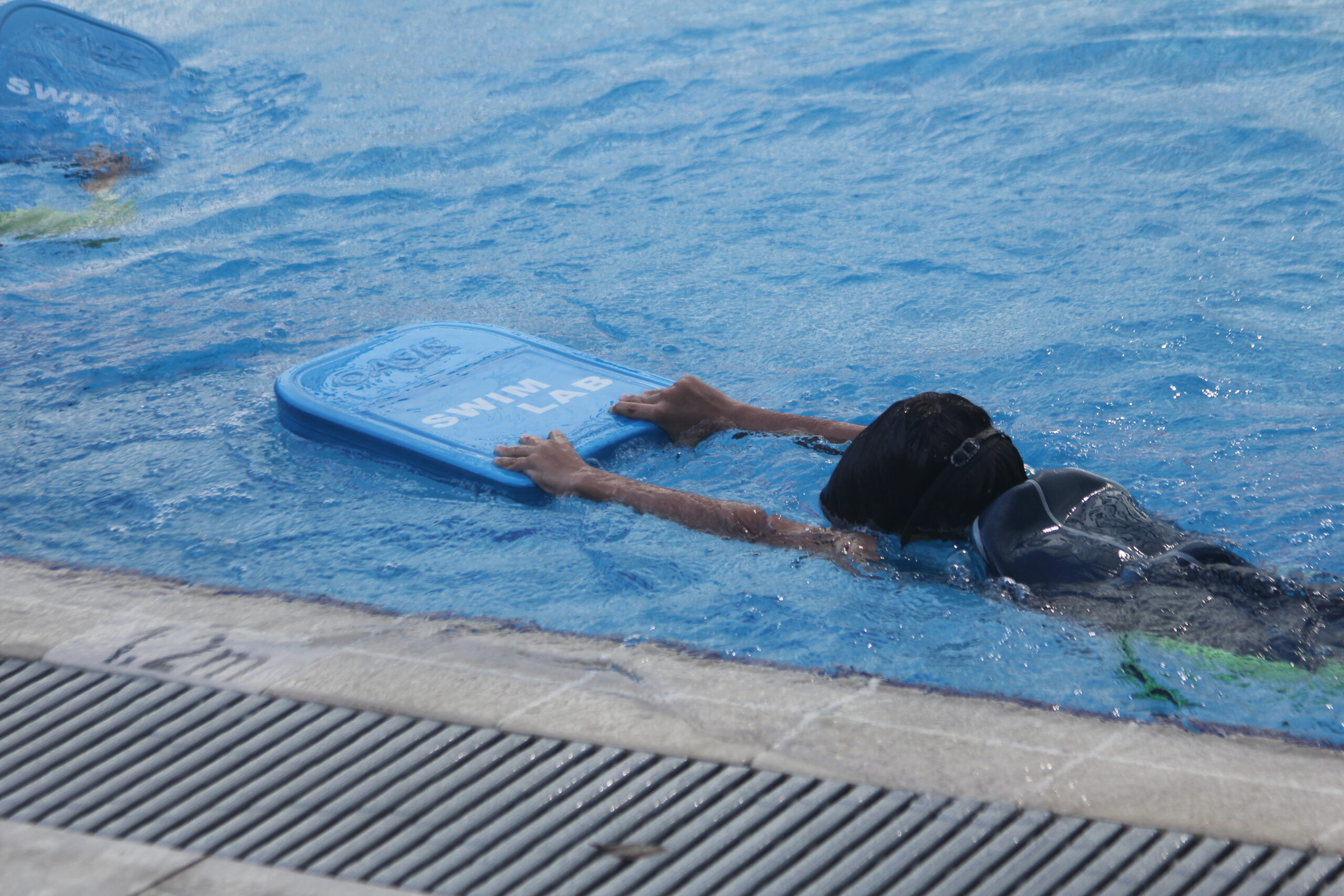 A child confidently swimming in The Swim Lab's pool using a float board with the 'Swim Lab' logo on it.
