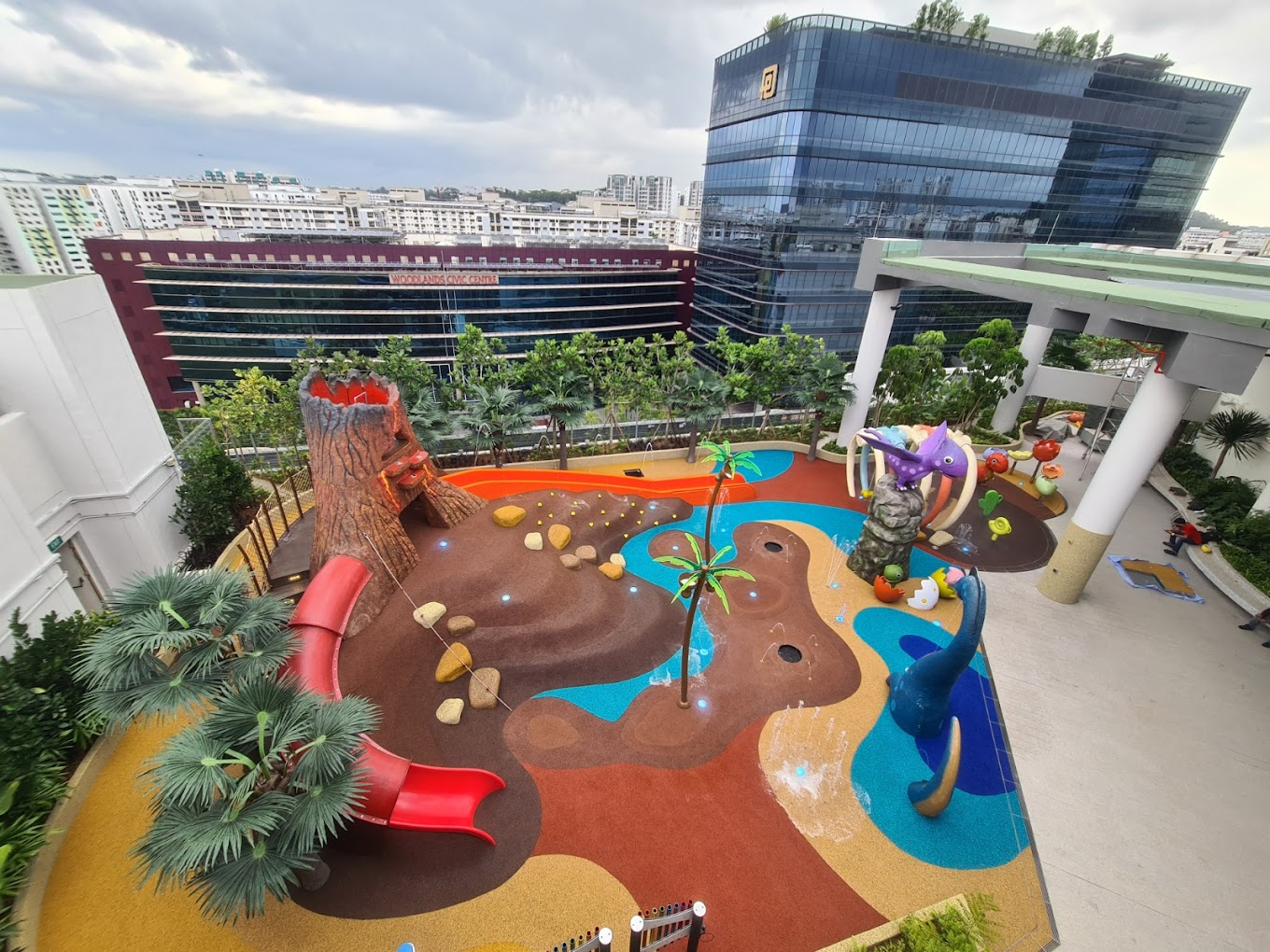 Water play zone adorned with dinosaur elements and a slide with a volcanic entrance, adjacent to Cantine by Kopitiam.