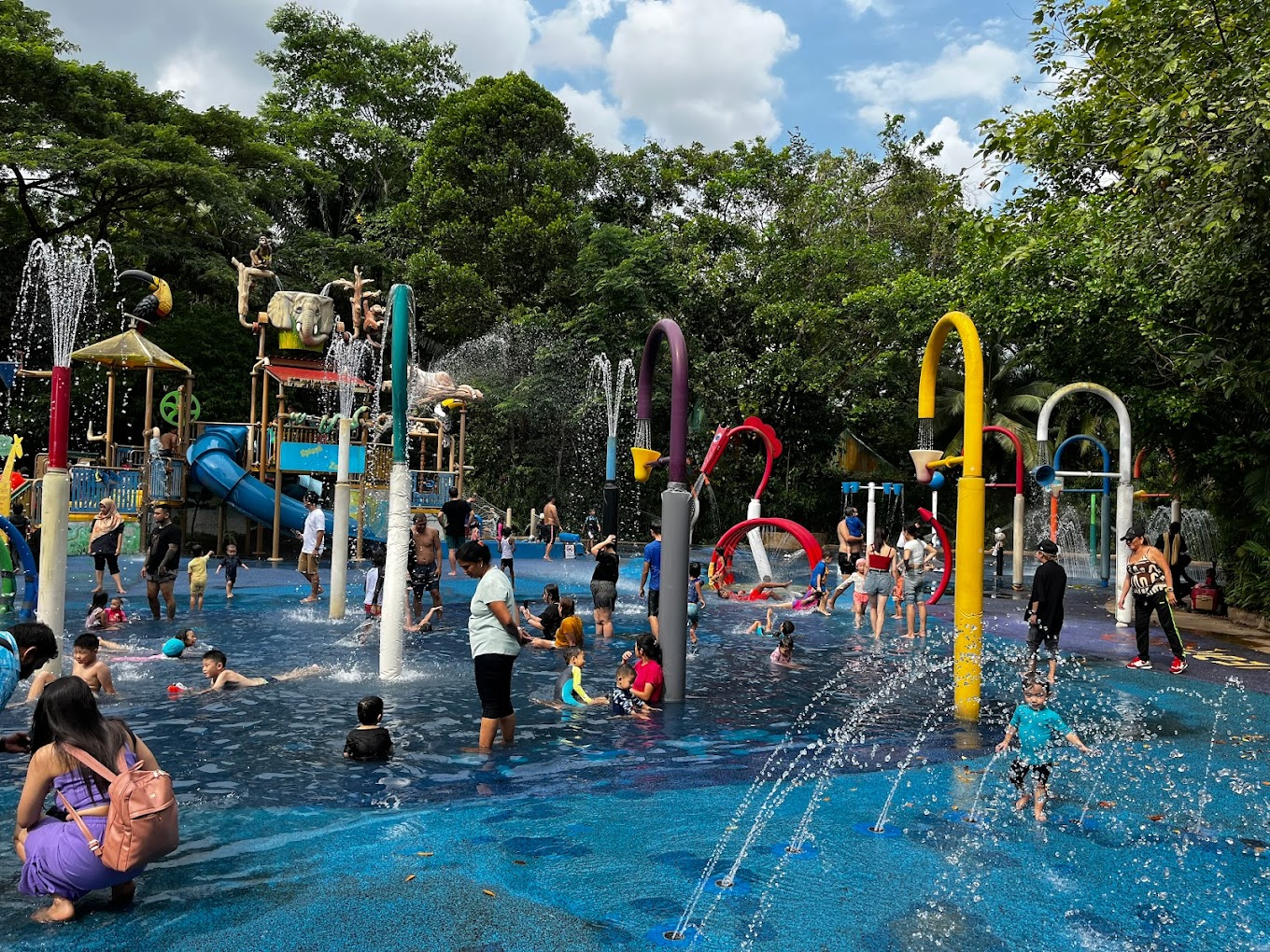 Water play area featuring splashing buckets, rain arches, water showers, and slides, located in Singapore Zoo with nearby food and beverage options.